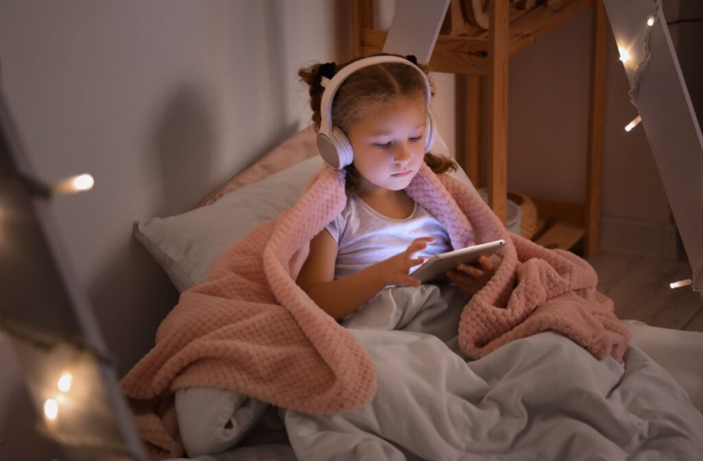 A young child wearing headphones and looking at a bright tablet in a dimly lit room